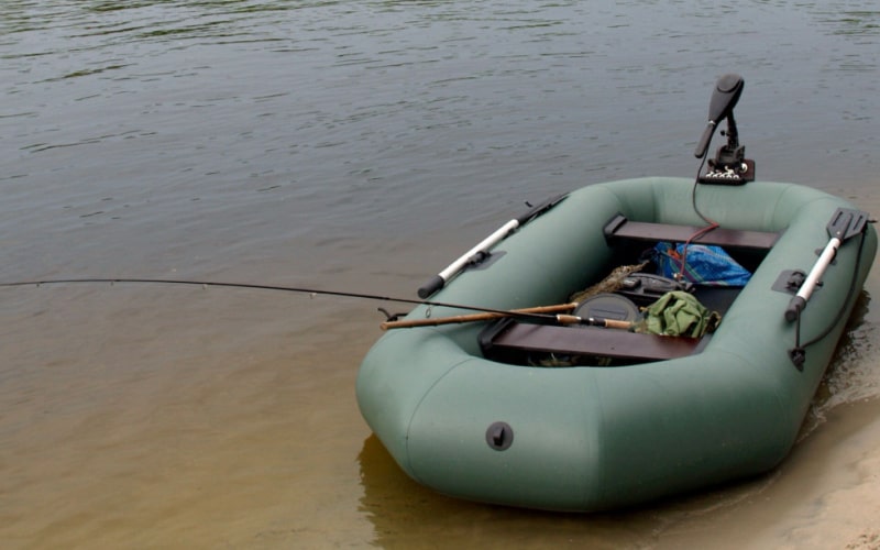 electric trolling motor fully connected to its battery is placed in a green inflatable boat on a riverbank in gloomy weather