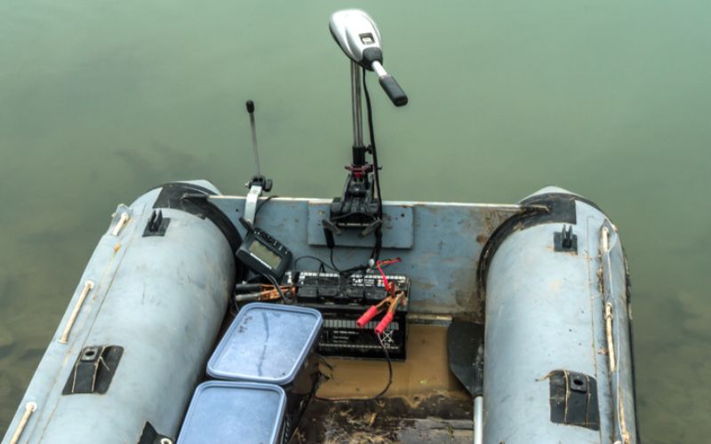 an old inflatable boat at river bank is equipped with a trolling motor connected to a marine battery